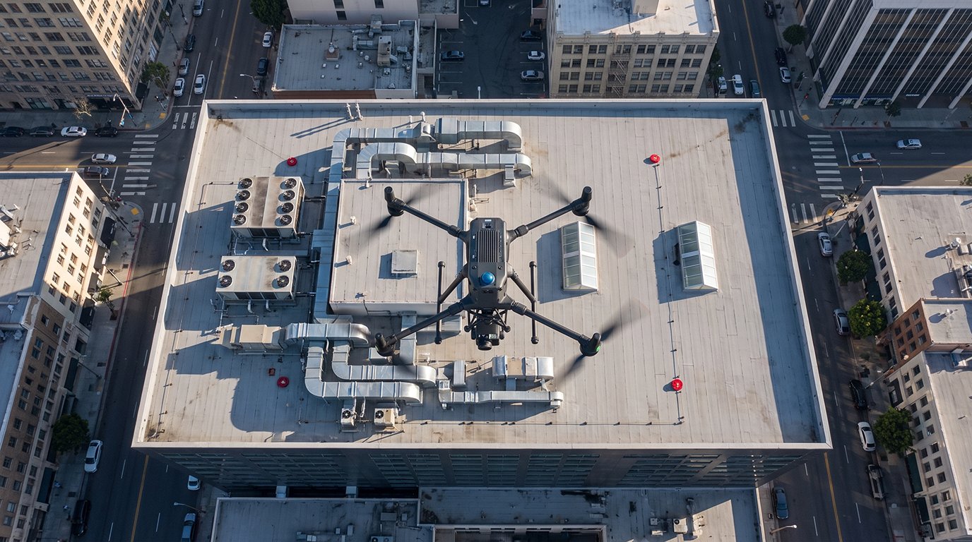 An aerial top-down view of an industrial inspection drone hovering over a flat city rooftop filled with complex HVAC ductwork and ventilation systems.